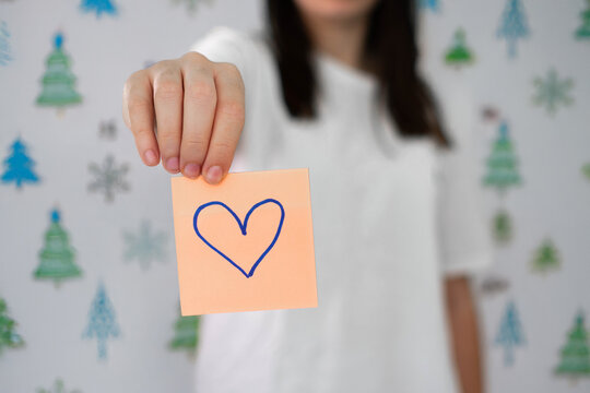 Close-up of a hand holding an orange sticky note with a blue heart drawn on it, symbolizing love, kindness, and emotional support.
