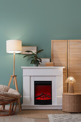 Fototapeta premium Interior of living room with folding screen, glowing lamps, armchair, wicker table and home decor on fireplace near blue wall