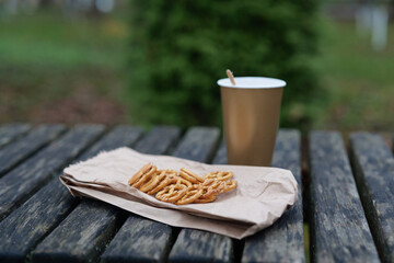 A warm drink sits beside a paper bag with pretzels, placed on a wooden table in a peaceful park setting during a cool afternoon