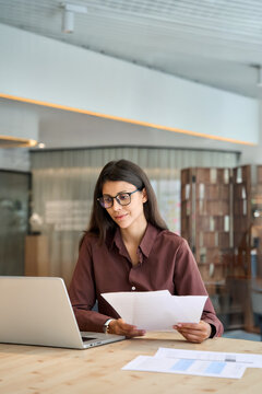 Concentrated latin business woman saleswoman working on laptop computer reading financial bank tax document in office. Young manager businesswoman specialist doing bill paperwork using pc. Vertical
