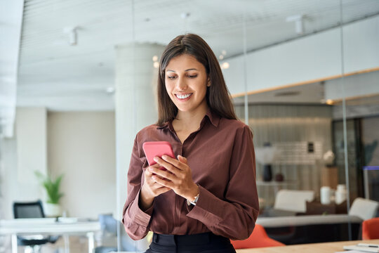 Young Indian business woman manager ceo user using cell phone mobile app. Smiling Latin Hispanic mature female businesswoman holding smartphone in office trading, reading working online at workspace