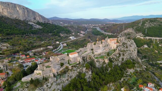 Aerial view of the historic Klis Fortress in Croatia, nestled amidst mountains and a vibrant village. Golden hour light illuminates this ancient stronghold and its picturesque surroundings.