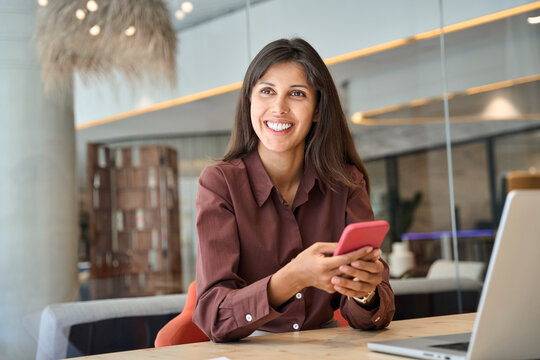 Hispanic business woman manager using cell phone mobile app, laptop. Smiling Latin or Indian student girl holding smartphone sitting in office or library, working online on gadget, dreaming smiling