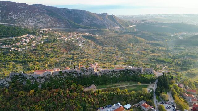 Aerial view of the historic Klis Fortress in Croatia, bathed in warm golden hour light, nestled beneath rugged mountains. A timeless blend of nature and heritage.