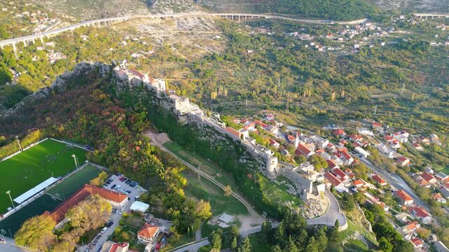 Aerial view of the historic Klis Fortress in Croatia, bathed in warm golden hour light, nestled beneath rugged mountains. A timeless blend of nature and heritage.