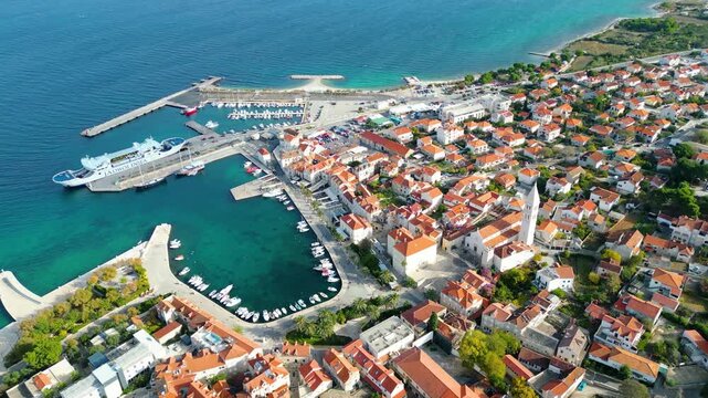 Aerial view of Postira, Brac Island, Croatia. The historic town, with its red roofs and bustling harbor, meets the clear blue Adriatic Sea, backed by distant mountains.