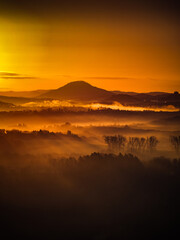 Vivid orange sunrise illuminating foggy lowlands and a distant mountain silhouette. Strong contrast, warm tones, and atmospheric depth create a dramatic and eye-catching landscape.