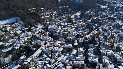 A beautiful aerial drone shot of Barzio blanketed in fresh winter snow, located in the Valsassina valley of Lombardy, northern Italy. The image highlights alpine homes, snowy landscapes, mountain ridg