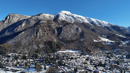 A beautiful aerial drone shot of Barzio blanketed in fresh winter snow, located in the Valsassina valley of Lombardy, northern Italy. The image highlights alpine homes, snowy landscapes, mountain ridg