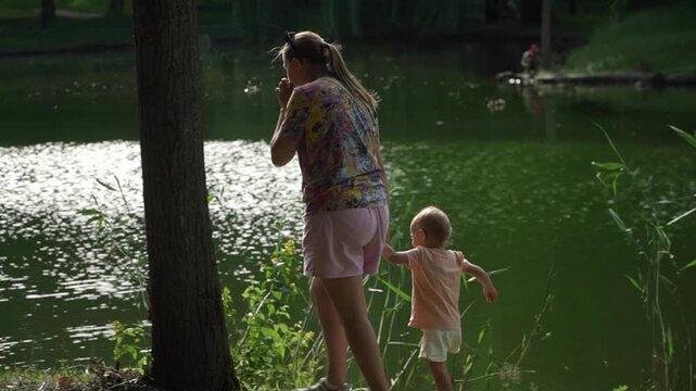 a girl with a baby approach the edge of the water of a beautiful green city lake in a park, looking at fish and birds, tall grass and reeds along the shore, a warm summer day