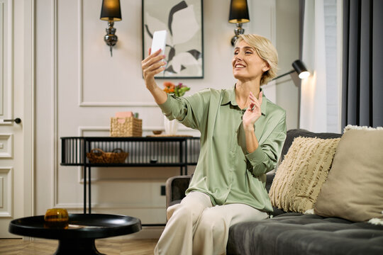 Caucasian middle aged woman sitting on sofa, smiling and waving while holding smartphone, taking selfie or video calling, in modern living room with decorative elements visible