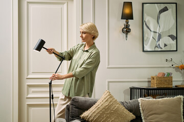 Middle aged Caucasian woman adjusting standing lamp in modern living room, standing near sofa and decorative pillows, focusing on arranging lighting fixture, neutral expression