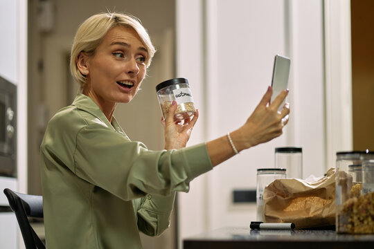 Caucasian young adult woman holding transparent food container while smiling and taking selfie with smartphone in kitchen, sitting at table with other containers and paper bag visible