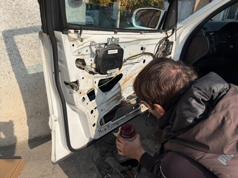 Auto mechanic or technician lubricating an open car door interior with red grease (lithium) during repair work. The car door's inner structure is exposed in the sunlight. Car service, repair, maintena