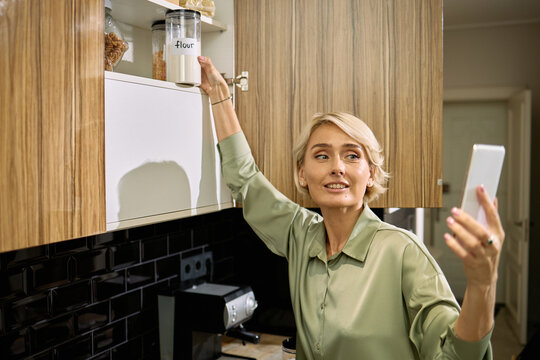 Caucasian middle aged woman reaching for flour jar on kitchen shelf while holding smartphone and smiling, multitasking during cooking or baking preparation in modern home kitchen