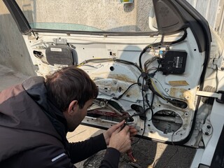 Auto technician working with hands on the mechanical and electrical components inside an open car door. The exposed door structure shows wires and metal parts. Car service, technical maintenance.