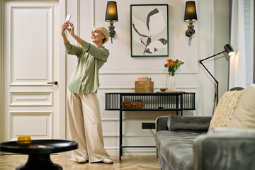 Caucasian senior woman smiling while taking selfie with smartphone standing in modern living room, holding phone at arm's length, enjoying moment, casual home environment