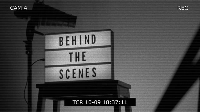 A cinematic view of a Behind the Scenes sign on a wooden stool under professional studio lighting. The frame includes a timestamp and recording elements mimicking a video camera interface. Close up.