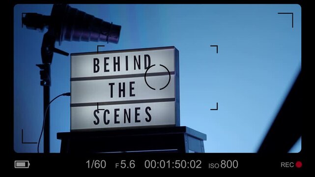 A cinematic view of a Behind the Scenes sign on a wooden stool under professional studio lighting. The frame includes a timestamp and recording elements mimicking a video camera interface. Close up.