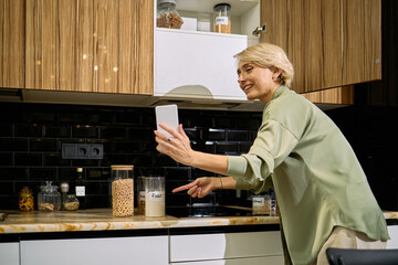 Caucasian middle aged woman smiling while holding smartphone and pointing at glass jar labeled flour in modern kitchen, engaging in video call or recording cooking tutorial