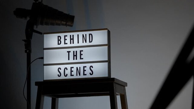 A cinematic view of a Behind the Scenes sign on a wooden stool under professional studio lighting. The frame includes a timestamp and recording elements mimicking a video camera interface. Close up.