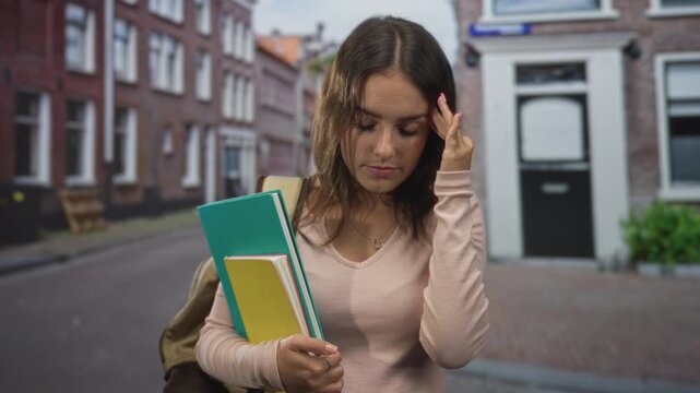 Woman holding notebooks and touching temple on street beside brick building and door, backpack slung over shoulder with green and yellow notebooks visible; academic stress.