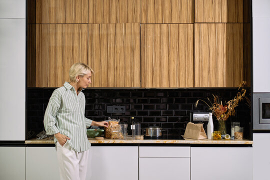 Caucasian middle aged woman standing in modern kitchen preparing breakfast with cereal and bread, looking at food on counter, kitchen appliances and dried flowers visible in background