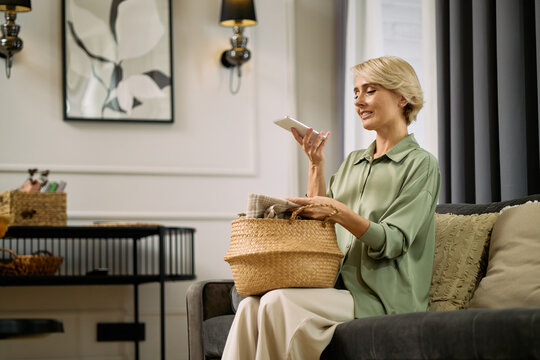Middle aged Caucasian woman sitting on sofa holding smartphone and speaking into device while packing woven basket, smiling and appearing engaged in multitasking at home