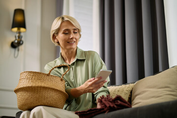 Caucasian middle aged woman sitting on sofa holding smartphone and woven basket, smiling while...
