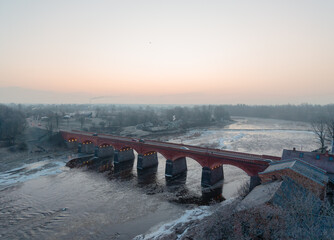 Aerial winter view of historic red brick bridge over partially frozen river with snowy banks and soft pastel sky, scenic European landscape background