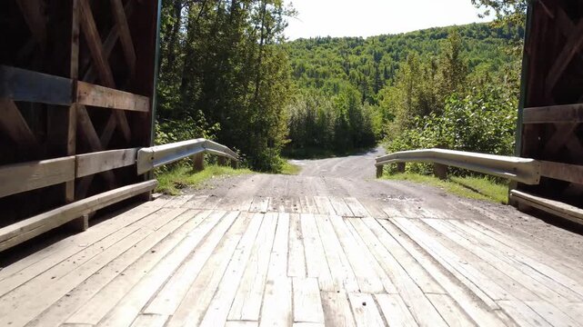 A quick passage through an old covered bridge, its structure entirely made of wooden planks and beams, leads to daylight and the forest at the exit. Belanger Bridge, Metis-sur-mer, Quebec, Canada.