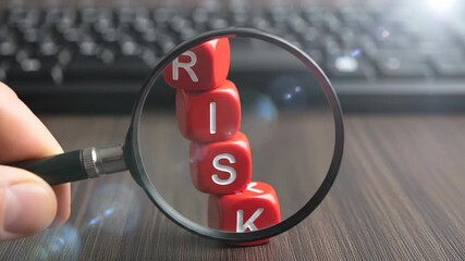 Hand holding magnifying glass focused on red wooden cubes spelling risk word on office desk near keyboard for business analysis and security assessment strategy management concepts