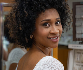 Close-up of a young woman with curly hair, dressed in white, looking back and smiling happily. You can see the reflection of a jewelry display case behind.
