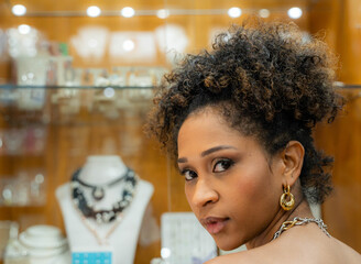 Close-up of a young African-American woman with curly hair and gold earrings, looking seductively over her shoulder. The blurred background is the display case of a jewelry store.