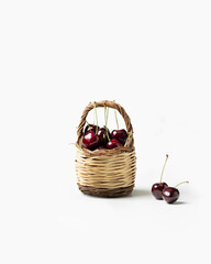 Fresh dark red cherries in a small woven basket with two cherries placed beside it on a clean white background, symbolizing summer fruit, freshness, and natural produce.