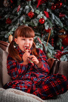 Portrait of a happy little girl child in pajamas sitting in christmas decorated room and licking a caramel glossy christmas candy cane lollipop. New Year's eve. St. Nicholas Day. Present. Holidays.	
