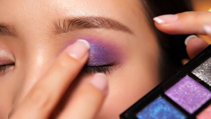 Close up shot of a skilled makeup artist expertly applying a bold and dazzling purple and blue shimmer eyeshadow onto a woman's eyelid using her fingertip