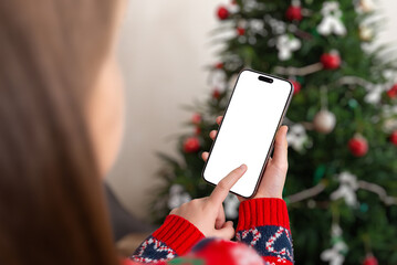 Girl holding smartphone with blank screen near Christmas tree, perfect for holiday app mockups,...