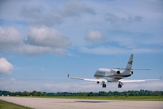 Rear view of a Gulfstream 260 beginning climb-out, wheels off the runway, wide angle view showing early ascent under bright daylight with soft clouds and tree line.