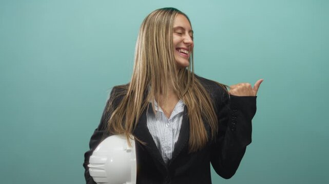 Smiling woman in a business blazer holding a white hardhat and pointing thumb over shoulder in a studio set; confidence ambition success.