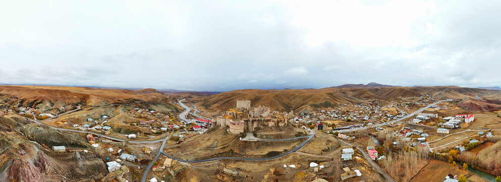 Panoramic aerial view of Hoşap Castle (Hoşap Kalesi) in Van, Turkey, majestically situated on a hill overlooking a scattered settlement and surrounding rugged, dry terrain under a dramatic sky. Travel