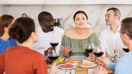 Joyful friends in their middle years sitting around table full of snacks and drinking wine in...