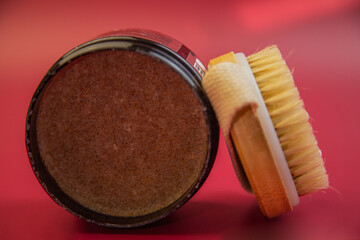Round container of body scrub positioned next to a wooden brush on a vivid red background, highlighting beauty and skincare essentials for pampering and self-care