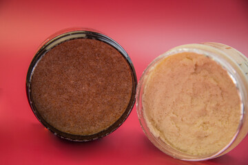 Two jars of body scrub, one brown and one beige, are displayed against a vivid red background, highlighting their unique textures and colors for skincare lovers