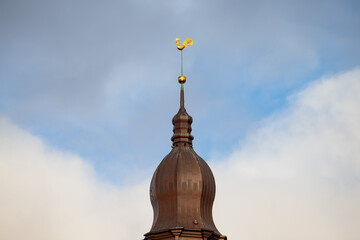 Tall spire in Riga rising from brown dome roof with distinct golden rooster element fixed to upper section and visible cloud formations in pale sky backdrop © K