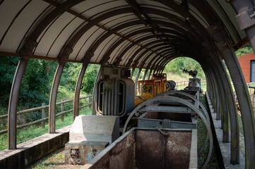 Row of retired mining equipment, including a metal tipping cart, power transformer, and industrial pump, housed in a curved, covered outdoor structure