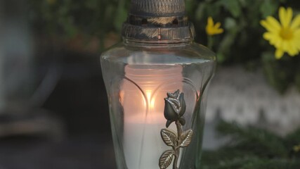 Close-up front view of a lit memorial candle on a grave, with a blurred background showing another candle and yellow flowers