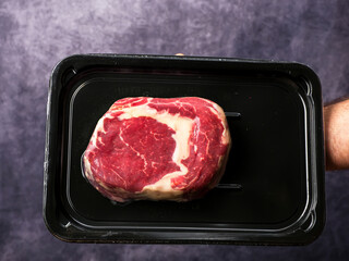 Premium top quality beef rib eye steak in black plastic vacuum packed package in butcher hand, painted grey background. Thick high value meat with lots of marbling for great taste and flavor.