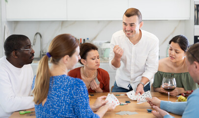 Excited man triumphantly displaying winning card combination during poker game with friends, during laid-back home gathering with wine around kitchen table