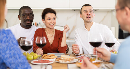 Casual evening where multiethnic group of cheerful carefree adult friends sharing wine, dining together, and chatting in cozy home kitchen
